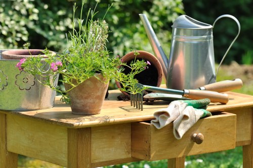 A close-up view of a garden wooden workbench set outdoors in a lush, green environment, featuring a variety of gardening tools and plants. Potted herbs with delicate green leaves and small purple flowers are placed on the workbench, alongside metal watering cans, garden forks, and a pair of work gloves. The workbench has a small drawer with a wooden knob, and the surface shows signs of recent use, with some soil and plant residues visible. The background includes a blurred garden scene with leafy shrubs and trees, suggesting a well-maintained outdoor space suitable for gardening and landscaping activities in Bethnal Green or nearby London areas. Natural daylight illuminates the scene, highlighting the textures of the wooden surface and greenery, creating a vibrant and professional gardening setting consistent with services provided by Hedge Trimming Bethnal Green.