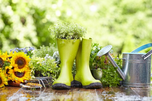 The image displays a gardening scene set outdoors with a blurred background of green foliage, indicating a garden environment. In the foreground, there is a wooden garden table with visible grain and a slightly weathered surface, supporting various gardening tools and decorative items. Prominently placed are a pair of bright green rubber gardening boots filled with white and green flowering plants, positioned upright and serving as unconventional planters. To the right of the boots, a traditional metal watering can with a long spout and a blue handle sits next to a small potted green shrub. On the left side of the table, vibrant yellow sunflowers with dark centers are arranged alongside others of different colours, enhancing the natural, lively atmosphere. The textures range from the smooth rubber boots and shiny metal watering can to the rough wooden surface of the table and textured plant leaves. The overall scene conveys an outdoor gardening setup, highlighting tools and decorative elements that complement gardening services in Bethnal Green, with the subtle presence of greenery and flowering plants creating a fresh, inviting outdoor space under natural lighting.