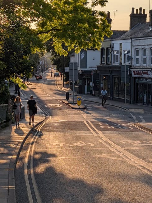A quiet street in Bethnal Green during late afternoon, featuring a row of traditional terraced houses with white and black facades on the right side, and lush green trees casting shadows on the asphal