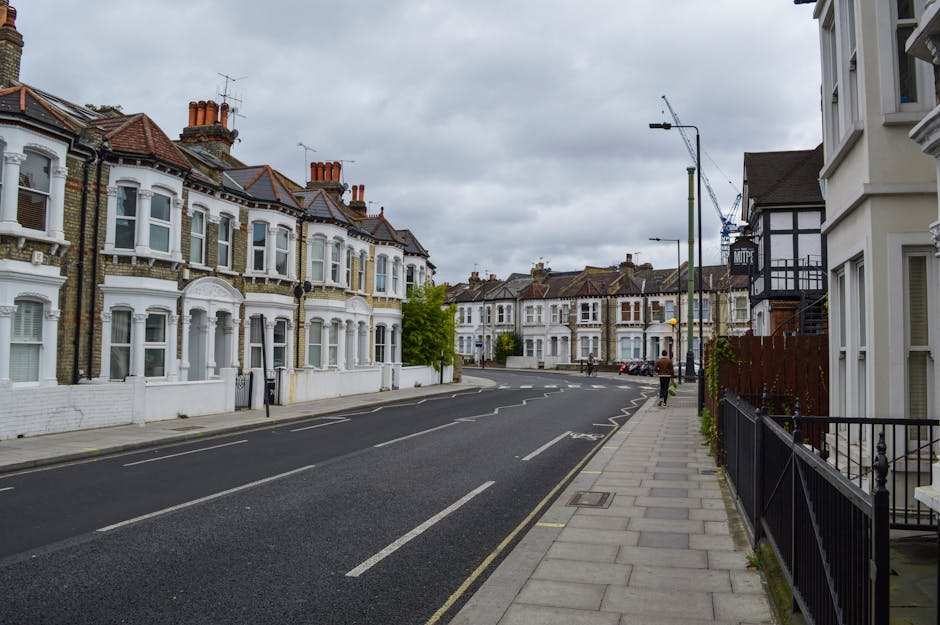 A quiet street in Bethnal Green during late afternoon, featuring a row of traditional terraced houses with white and black facades on the right side, and lush green trees casting shadows on the asphal