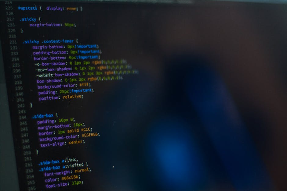 The image shows a close-up of a computer screen displaying lines of coloured programming code with syntax highlighting in green, blue, purple, and orange on a black background. The text appears slight